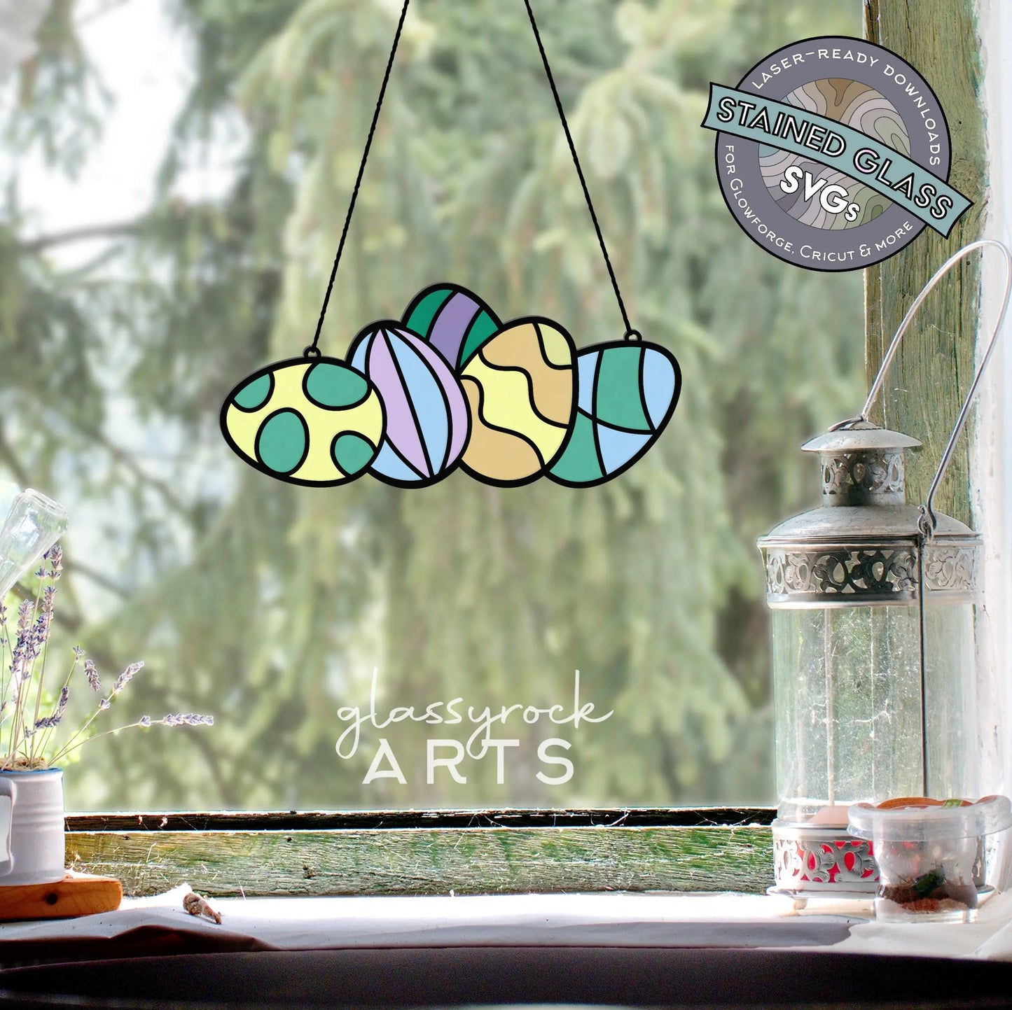 A stained glass Easter egg decoration with colorful abstract shapes hangs in front of a window, beautifully framed by a lush green view. On the ledge sit a lantern and potted plant. Text reads: glassyrock ARTS and Stained Glass Easter Eggs SVGs.