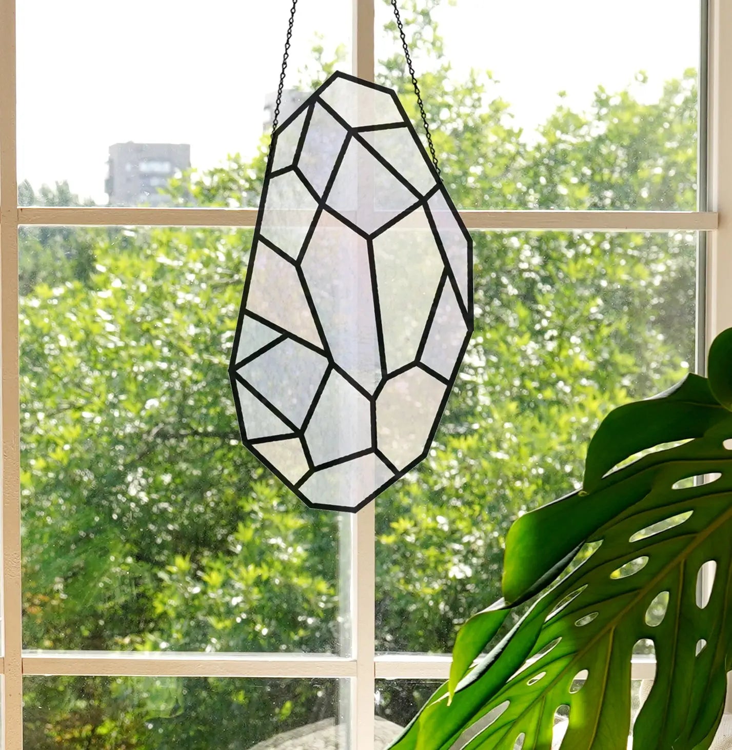 A Free Giant Gem from Noelle Barnes/GlassyRock Arts hangs in front of a window, revealing a lush tree outside. Sunlight casts intricate patterns through the glass. A houseplant with large leaves peeks into view at the bottom right. (Use code from your email at checkout.).
