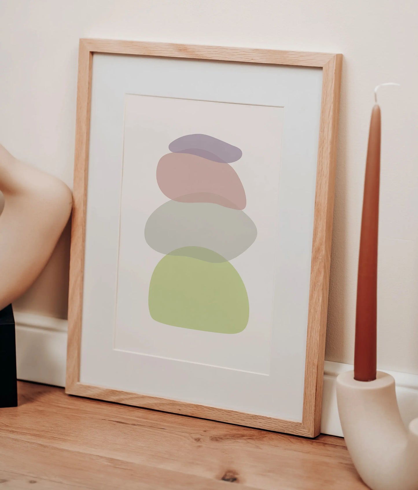 The Pastel Clear Modern Stone Shapes Digital Art leans against a cream wall on a wooden floor, accompanied by a tall brown candle in a white holder, creating a chic boho décor vibe.