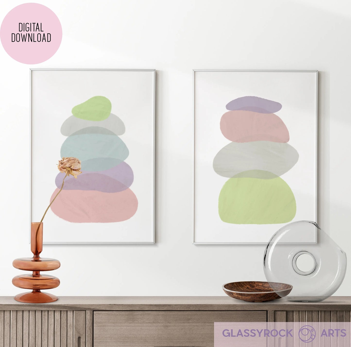 Two Pastel Painted Boho Stone Shapes Digital Art prints with multicolored ovals adorn the wall above a wooden sideboard. Emphasizing the boho theme, a dried flower in a modern glass vase and a glass ornament rest on the sideboard.