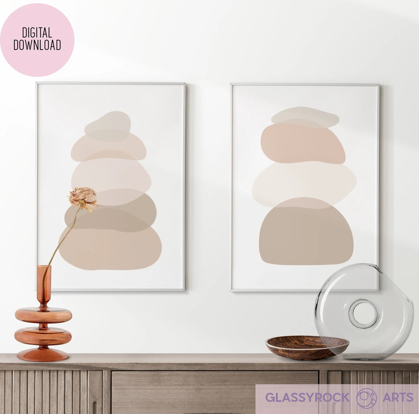 Two framed digital prints featuring Ombre Nude Stone Shapes with abstract rounded shapes in neutral tones adorn a white wall. Below, a wooden cabinet holds a modern amber vase with dried flowers, a clear glass decorative object, and a wooden bowl.