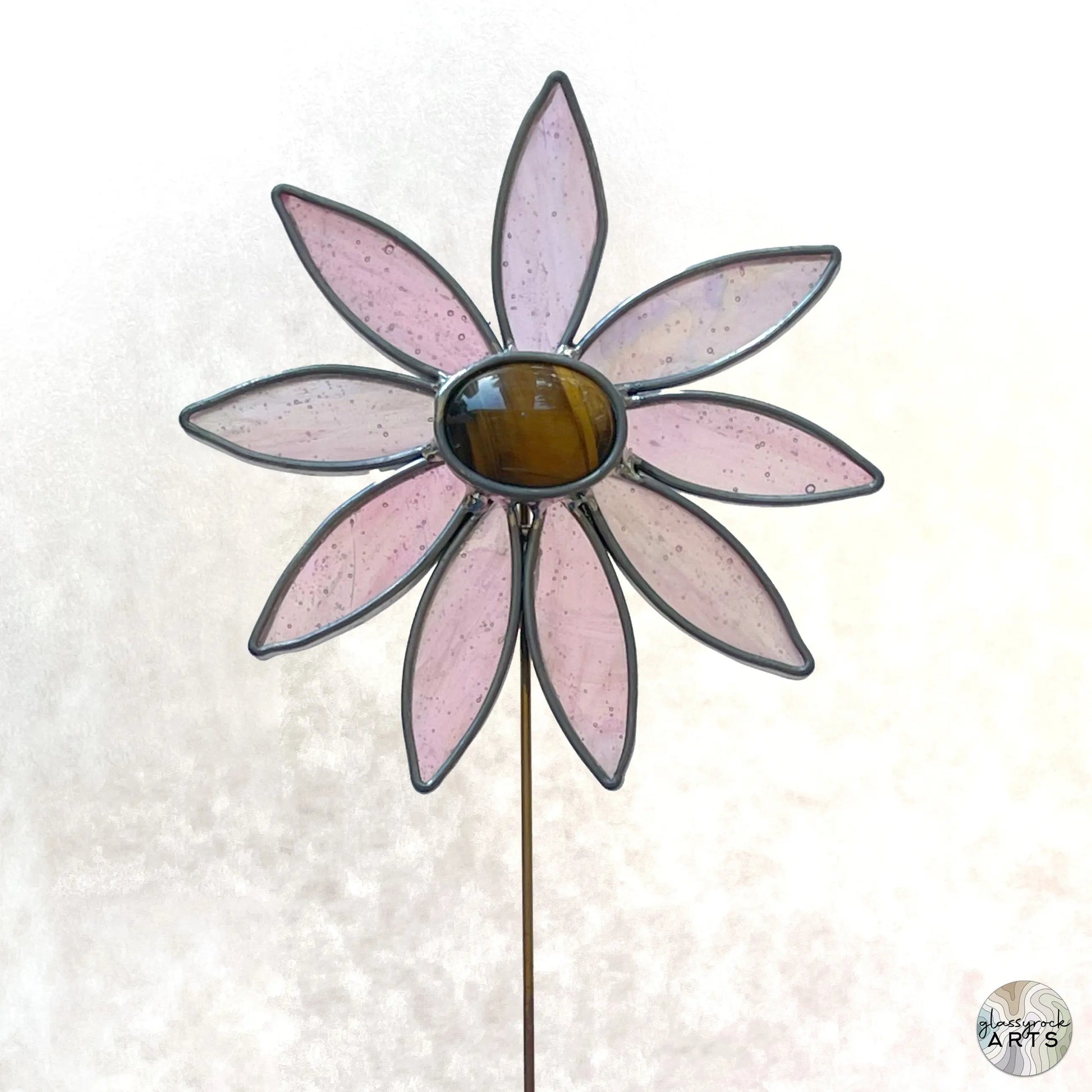 The Handmade Stained Glass Flower Plant Stake with Tigers Eye Center features pink petal-shaped glass and a brown center, using Tiffany stained glass methods. The flower is mounted on a thin rod against a light, textured background.
