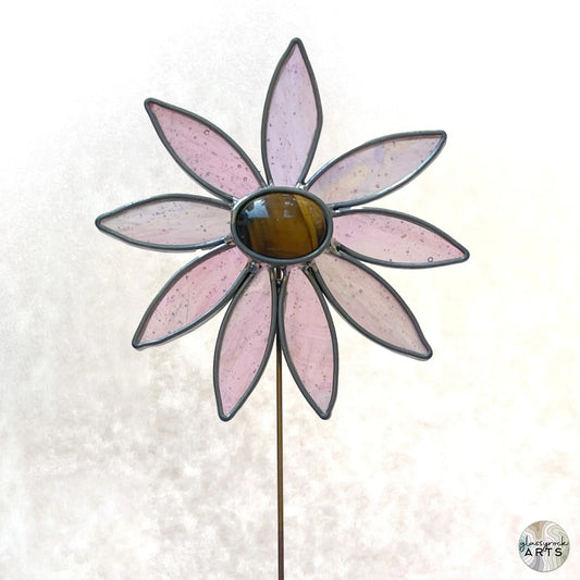 The Handmade Stained Glass Flower Plant Stake with Tigers Eye Center features pink petal-shaped glass and a brown center, using Tiffany stained glass methods. The flower is mounted on a thin rod against a light, textured background.