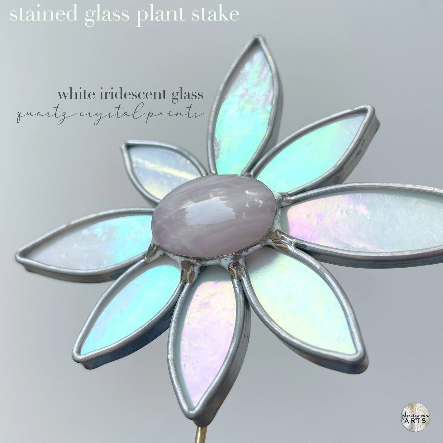 The Handmade Stained Glass Flower Plant Stake with Rose Quartz showcases white iridescent petals and a central quartz crystal on a light gray background. Crafted using Tiffany methods, this piece elegantly merges art with nature.