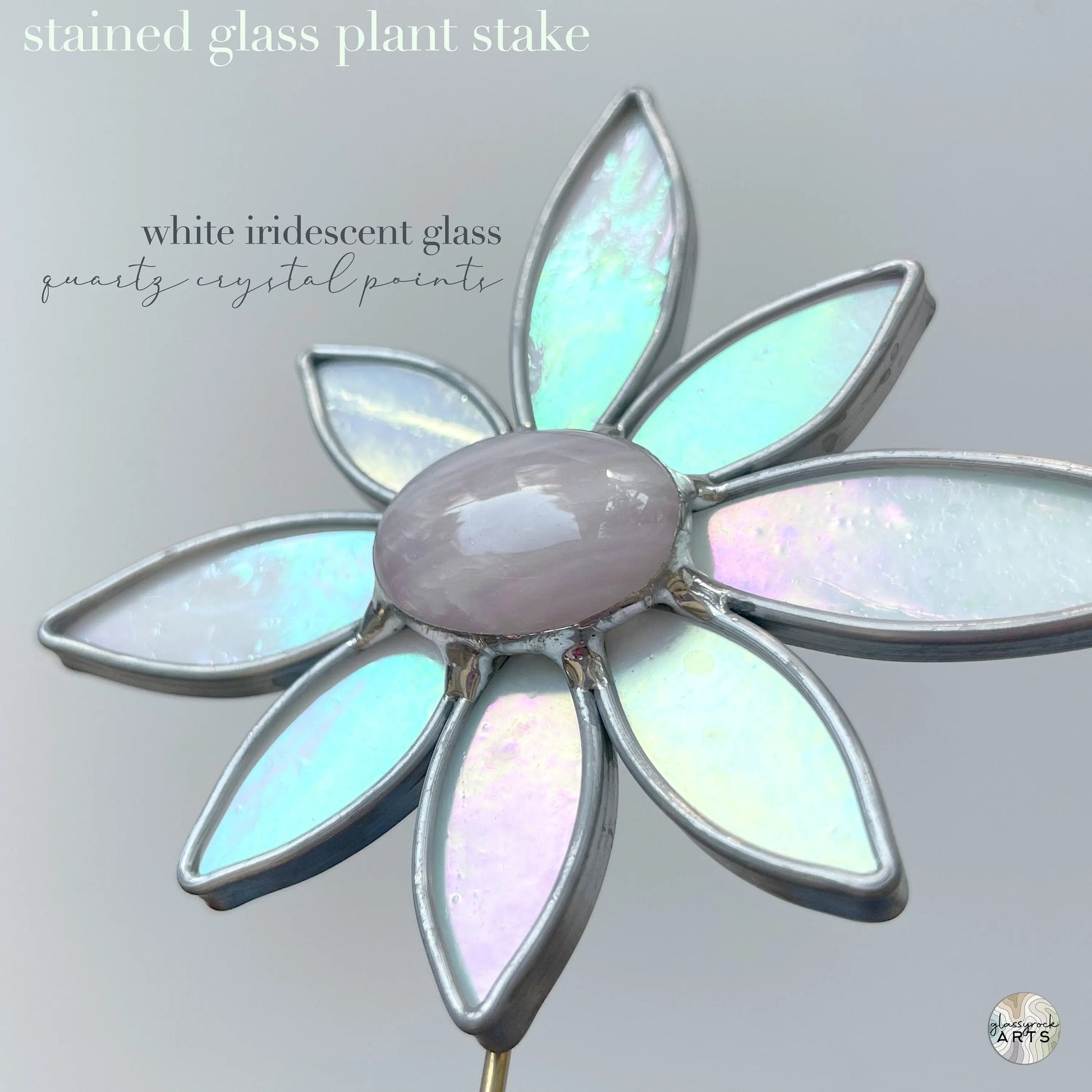 The Handmade Stained Glass Flower Plant Stake with Rose Quartz showcases white iridescent petals and a central quartz crystal on a light gray background. Crafted using Tiffany methods, this piece elegantly merges art with nature.