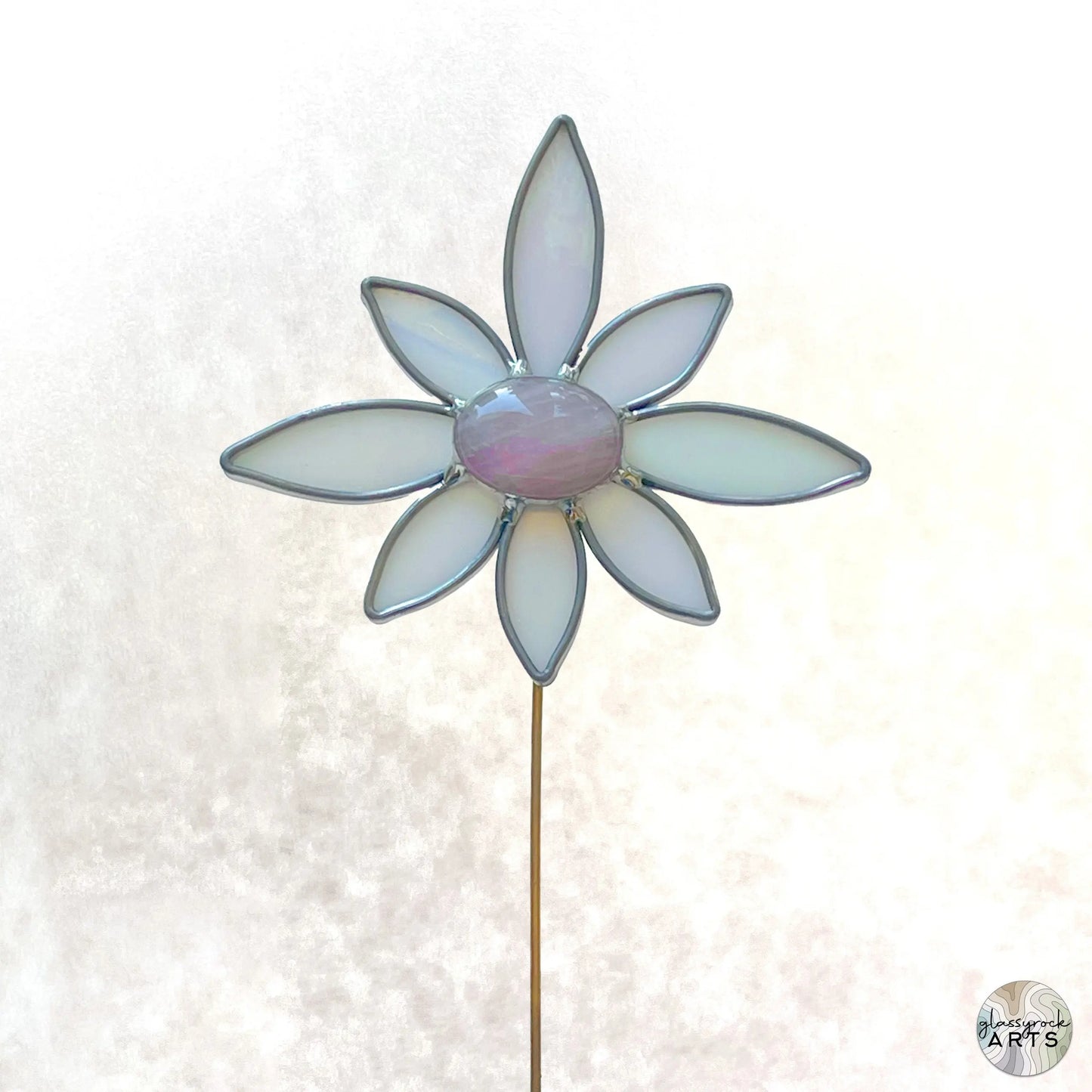 The Handmade Stained Glass Flower Plant Stake with Rose Quartz features seven white petals and a central pink piece, all edged in silver on a thin rod, resembling a delicate plant stake against a soft, blurred white background.
