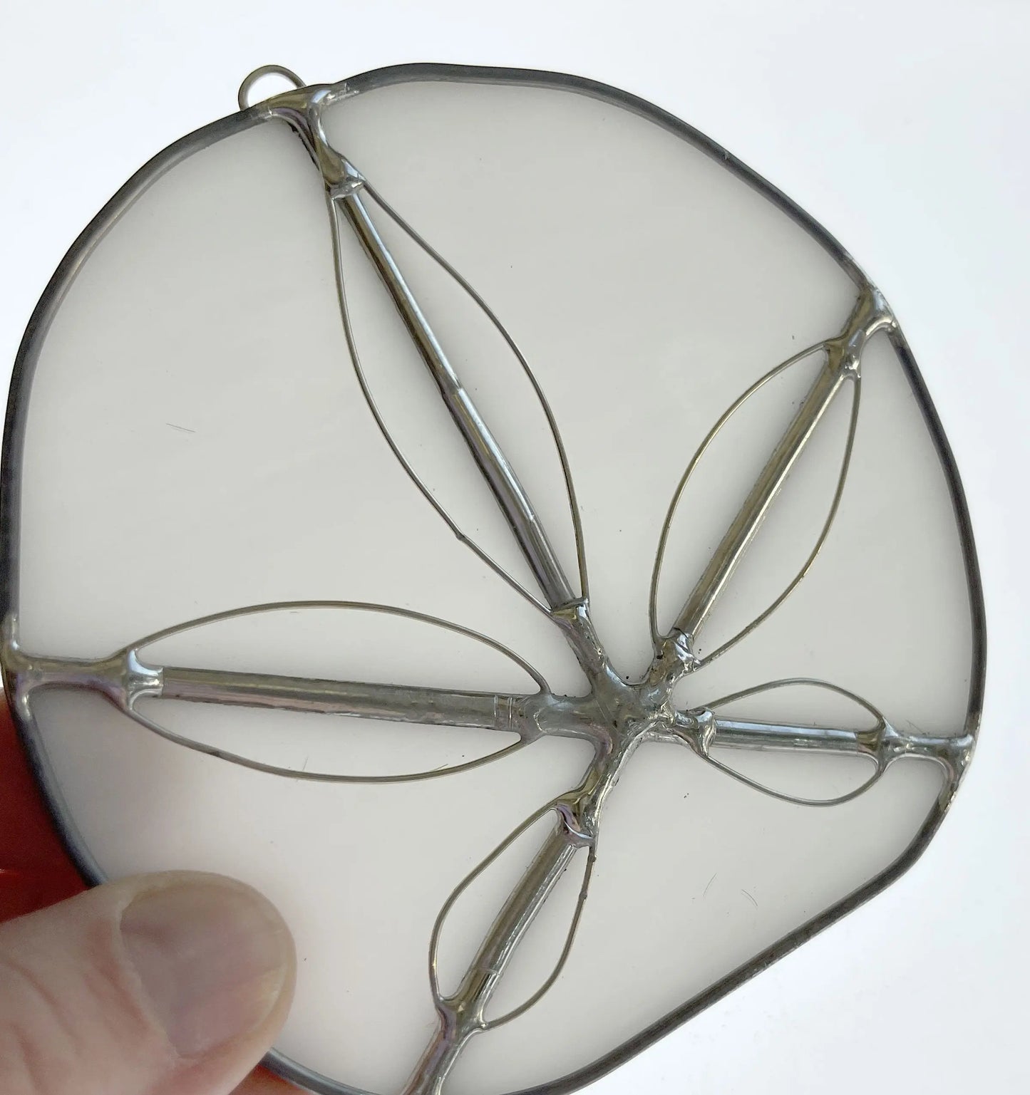 A hand displays a Handmade Stained Glass Sand Dollar - White, featuring translucent milky glass with a silver outline. Crafted by a Pacific Northwest artist, it resembles petal-like shapes in the sand dollar design, perfect as an elegant suncatcher.