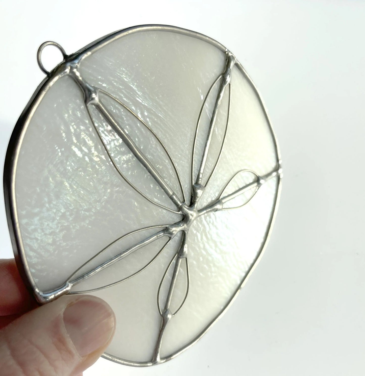 A hand holds a Handmade Stained Glass Sand Dollar, a circular ornament with a delicate silver frame. Crafted by a Pacific Northwest artist, its translucent white with a symmetrical leaf-like design. The plain white background highlights its handmade elegance.