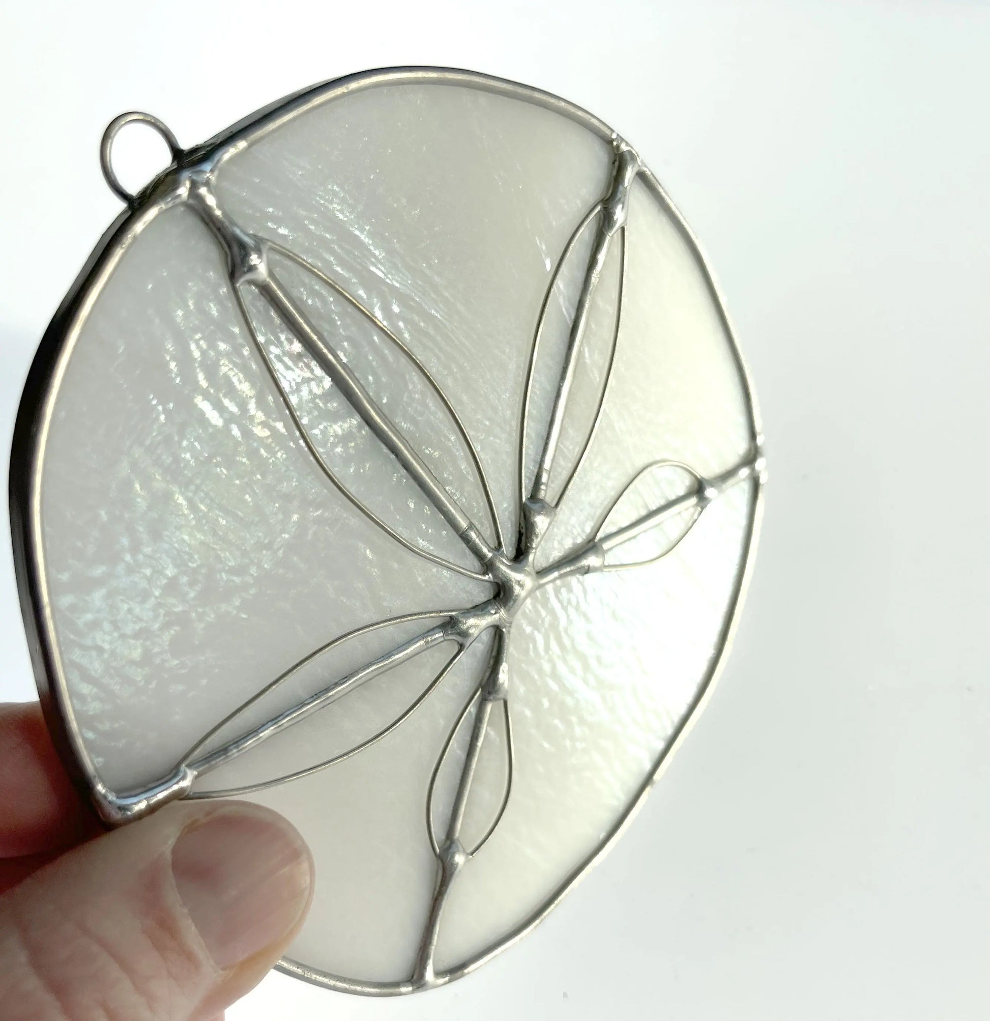 A hand holds a Handmade Stained Glass Sand Dollar, a circular ornament with a delicate silver frame. Crafted by a Pacific Northwest artist, its translucent white with a symmetrical leaf-like design. The plain white background highlights its handmade elegance.