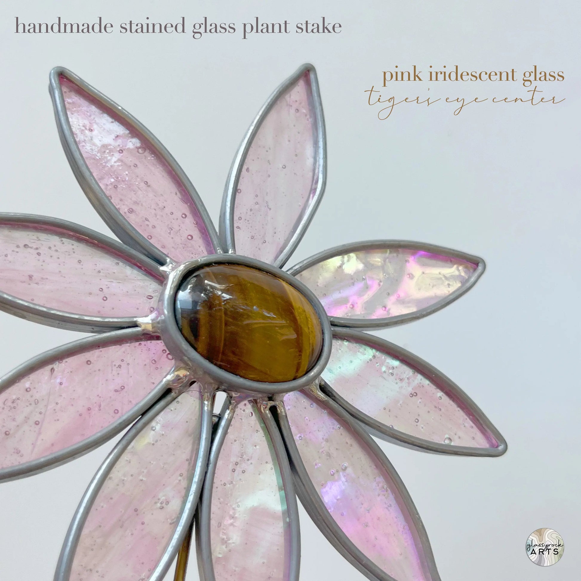 Crafted with Tiffany methods, this stained glass flower plant stake features pink iridescent petals and a tigers eye gemstone center, adding an artistic touch. Text in the image: handmade stained glass plant stake and pink iridescent glass tigers eye center.