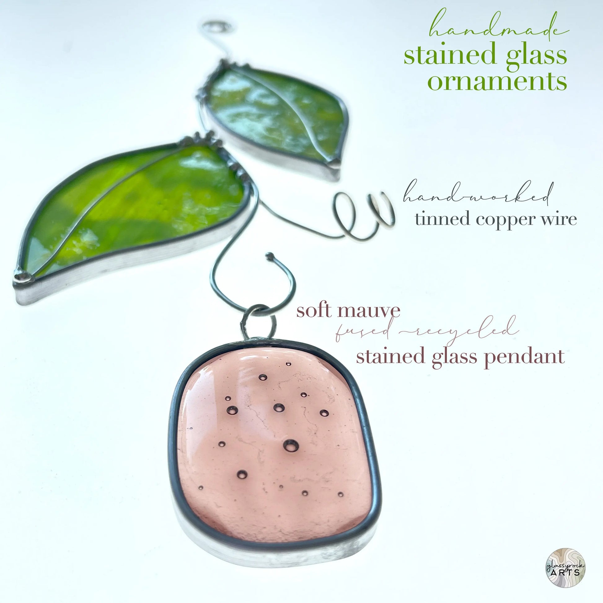 The Handmade Stained Glass Leaf Suncatchers set includes a soft mauve fused recycled glass pendant and green leaf-shaped pieces, linked with tinned copper wire, showcasing the beauty of handcrafted and upcycled stained glass.