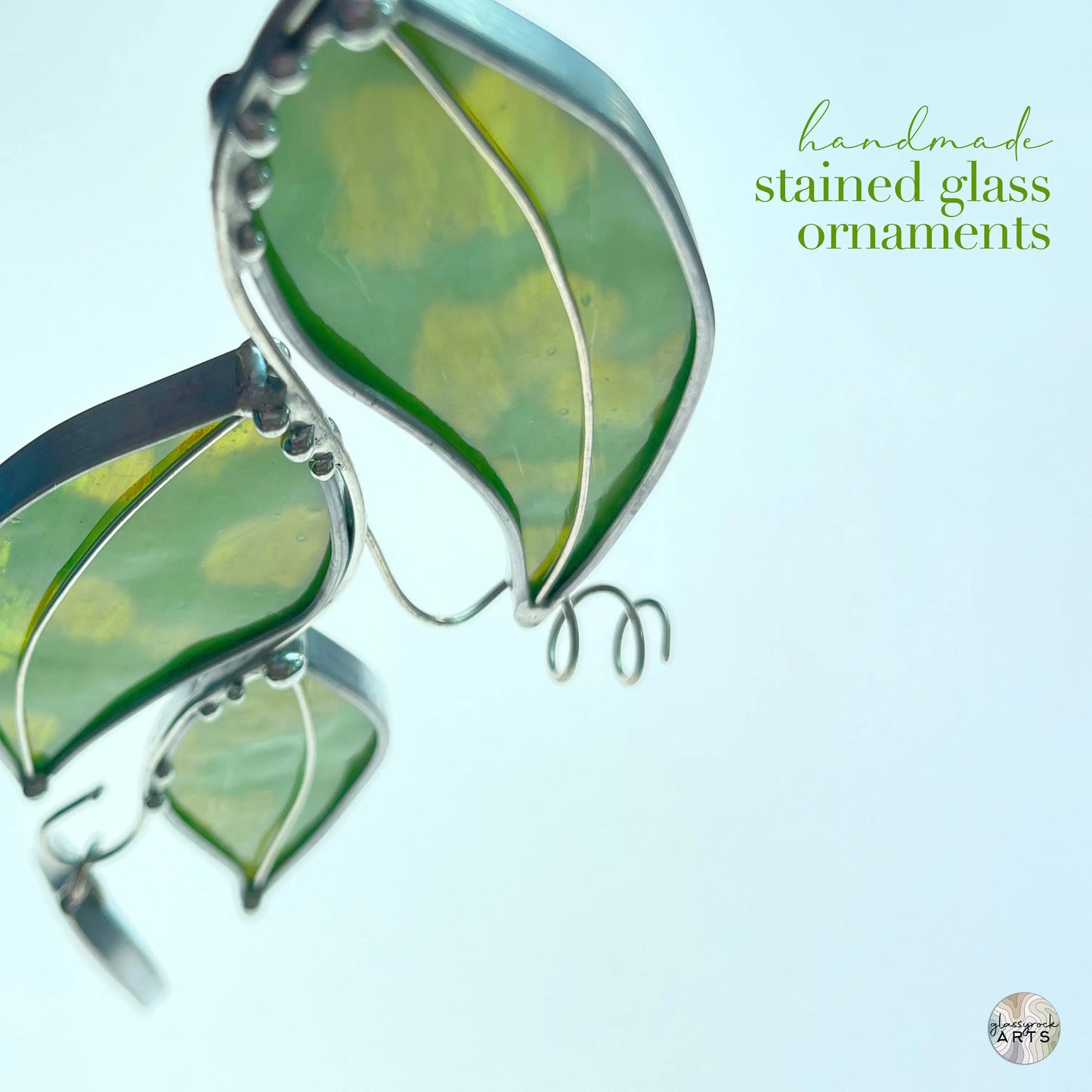 Close-up of three Handmade Stained Glass Leaf Suncatchers with green leaf designs, crafted using the Tiffany-method. Metal outlines accentuate the leaves against a soft white background. The text reads handmade stained glass ornaments, and a small logo appears in the corner.