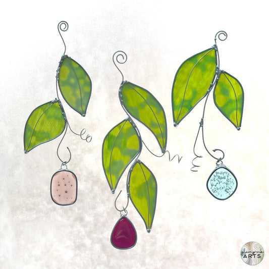The Handmade Stained Glass Leaf Suncatchers feature three metal-outlined leaves, each with a colored stone—pink, blue, and red. They dangle elegantly against a light background, giving off a whimsical and delicate look.