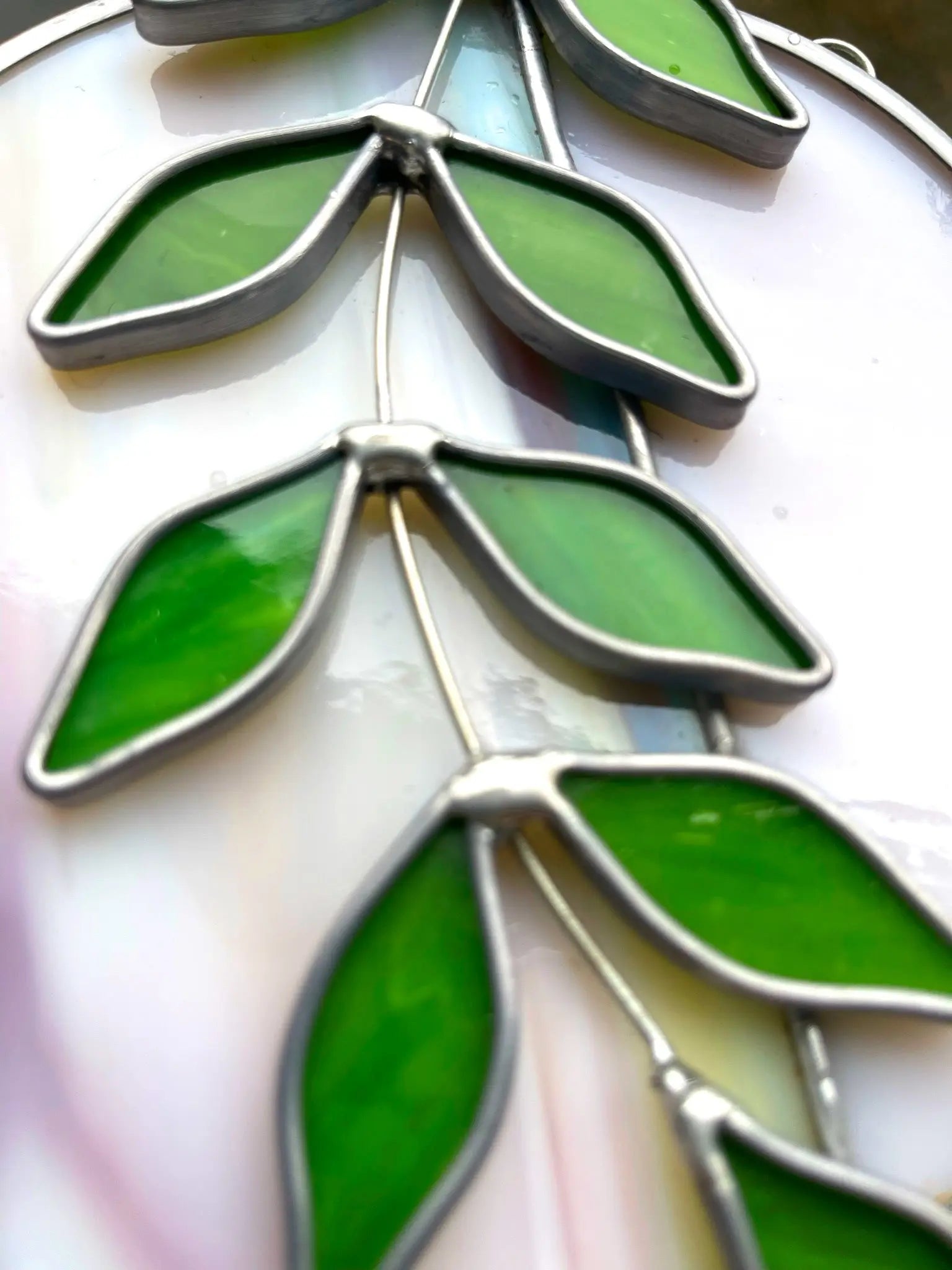 A close-up of the Handmade Ivy Rainbow Moon Stained Glass Suncatcher showcases a branch with green leaves outlined in metallic frames, set against a soft, muted color background.