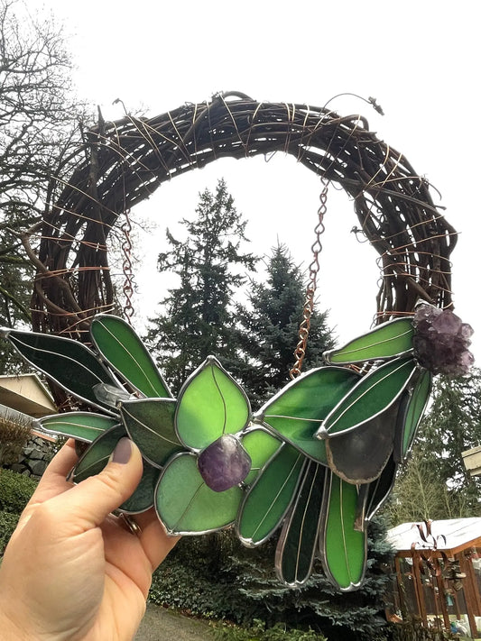 A hand displays the Handmade Botanical Stained Glass Wreath, featuring intertwined branches with green and purple glass resembling leaves. Trees and a cloudy sky in the background enhance its botanical artistry.