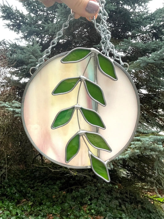 A hand delicately holds Ivy Botanicals Handmade Ivy Rainbow Moon Stained Glass Suncatcher, showcasing a branch with green leaves. The soft pink backdrop contrasts with the outdoor scene of evergreen trees and foliage framing this captivating stained glass artwork.