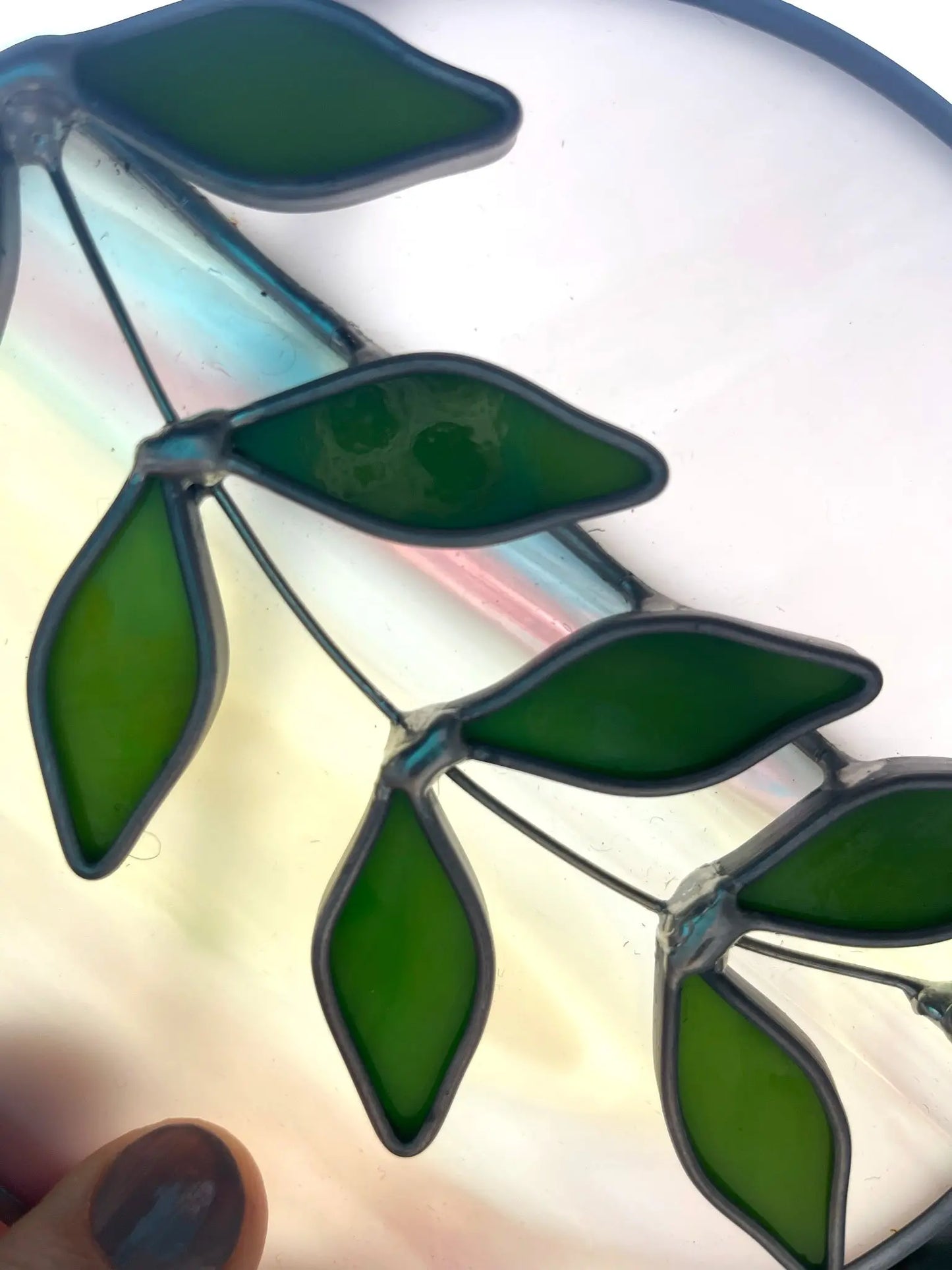 Close-up of the Handmade Ivy Rainbow Moon Stained Glass Suncatcher featuring green leaves on a translucent background. This art piece showcases leaves bordered by metal, with light streaming through to highlight its vibrant colors.