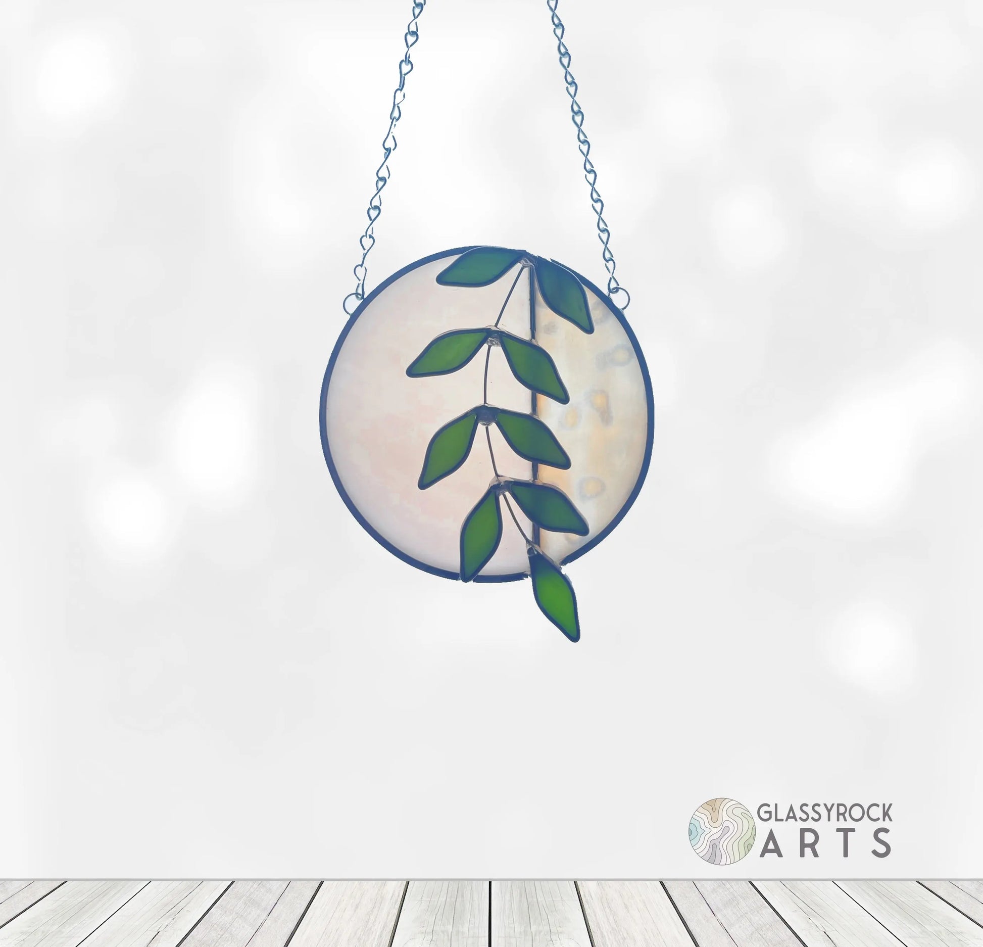 The Handmade Ivy Rainbow Moon Stained Glass Suncatcher, featuring a green leaf design similar to Ivy Botanicals, hangs elegantly from a chain. The background is softly blurred with a light wooden floor, and the Glassyrock Arts logo is in the corner.