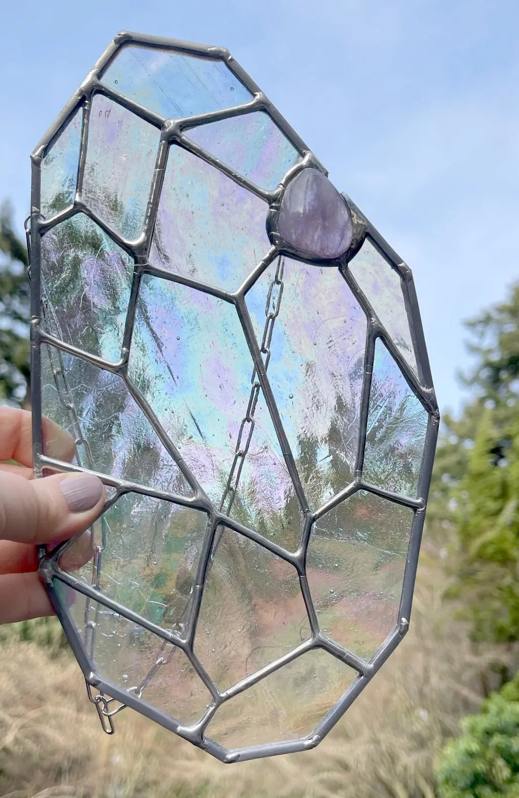An iridescent stained glass suncatcher, featuring a giant gem pattern and a purple stone accent, is held against a blurry backdrop of trees and clear sky, showcasing the unique artistry of Noelle Barnes/GlassyRock Arts. Try Free Giant Gem at checkout with your code.