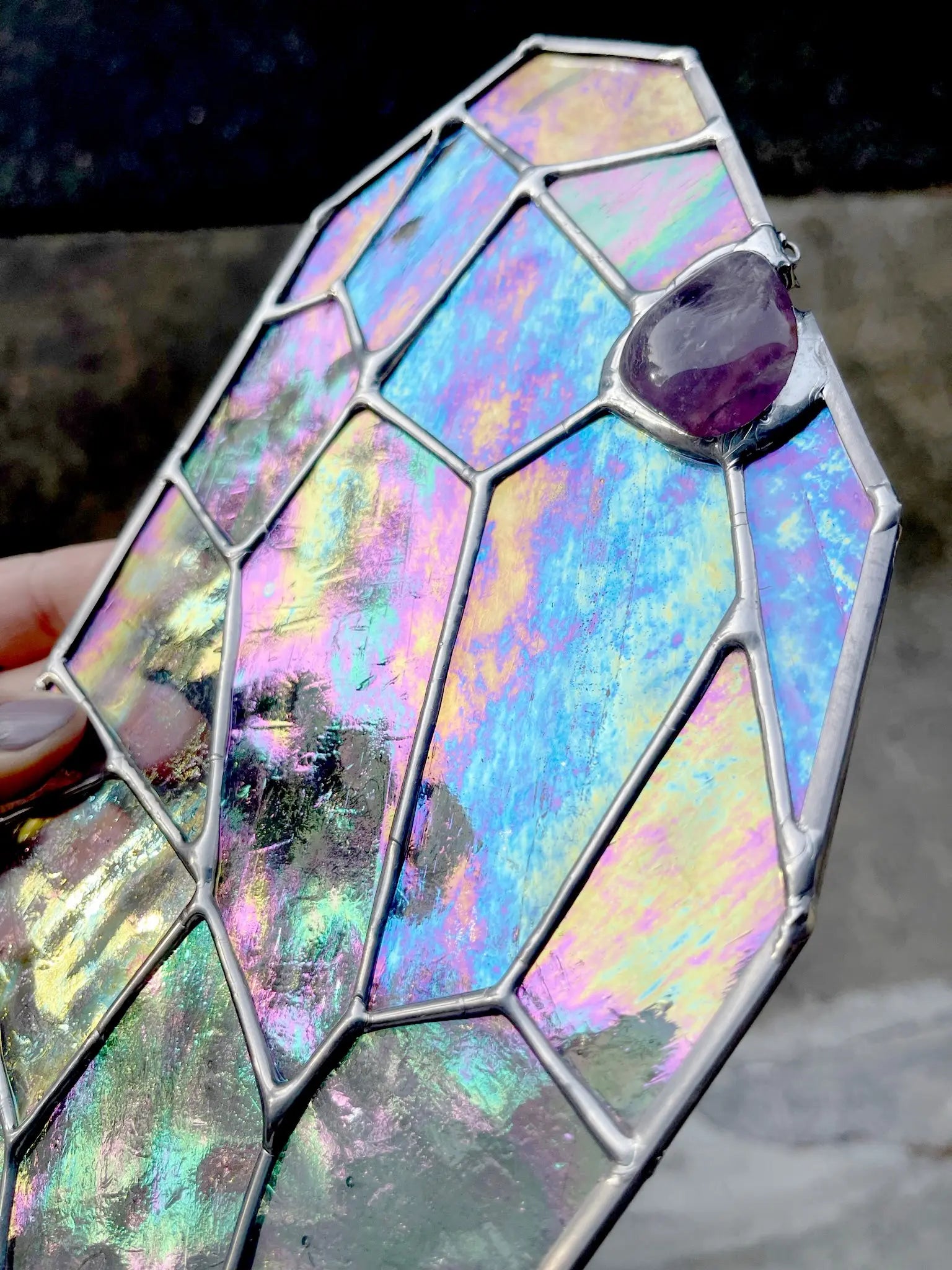 Close-up of a hand holding the Free Giant Gem glass piece with a geometric design in iridescent hues of blue, purple, and yellow. Adorned with a small purple stone at the top, this GlassyRock Arts creation by Noelle Barnes is set against a softly blurred outdoor background.