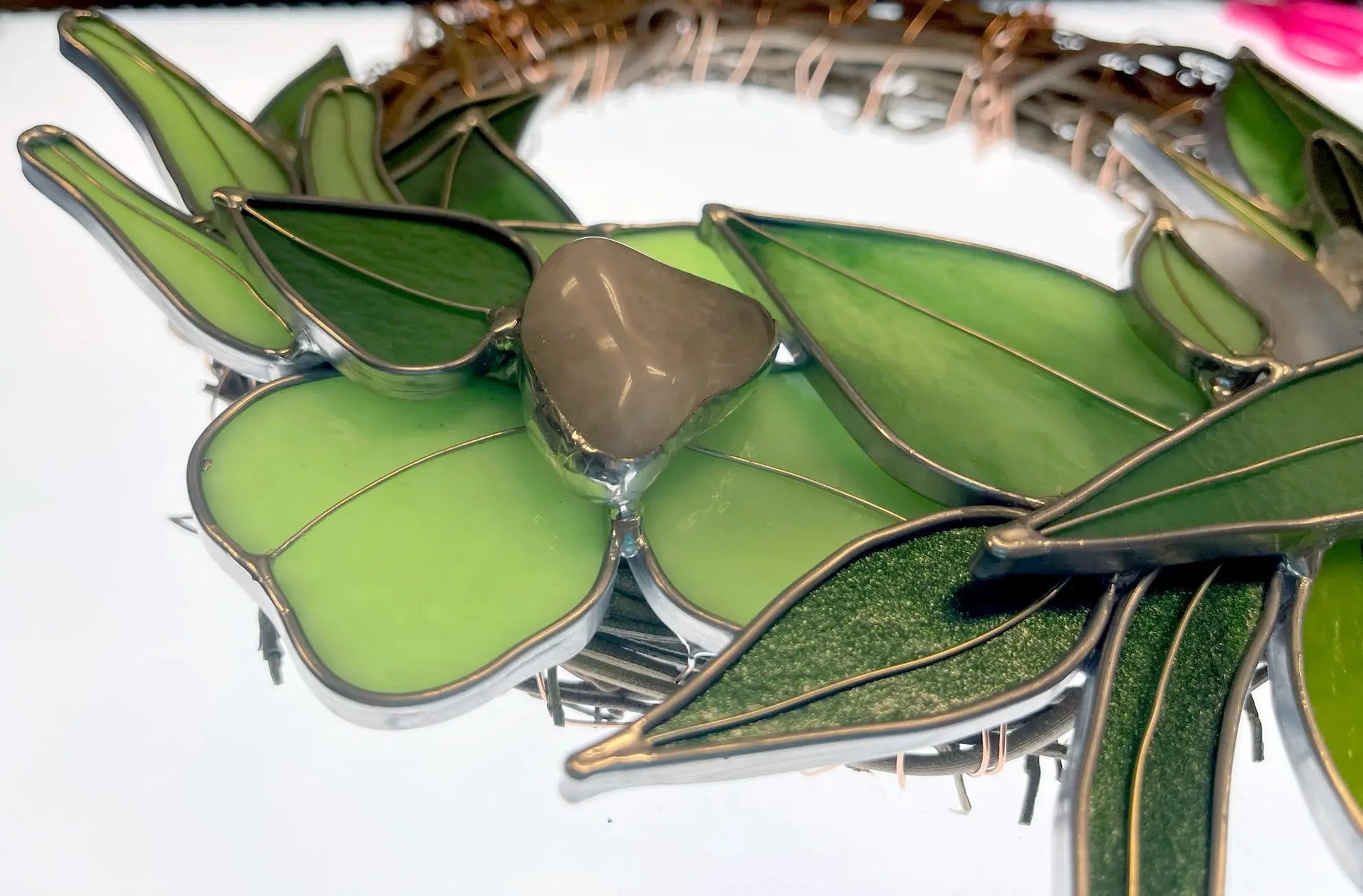 A close-up of the Handmade Botanical Stained Glass Wreath showcases green leaves with metal outlines and a central frosted gray stone, set against a softly lit background.