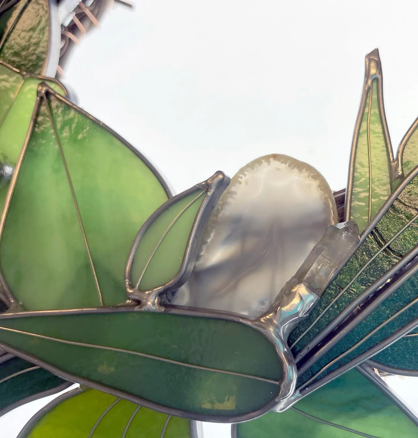 The Handmade Botanical Stained Glass Wreath showcases green leaves with copper outlines and a central white flower. Crafted from varying shades of green and translucent white glass, it elegantly captures natures beauty.