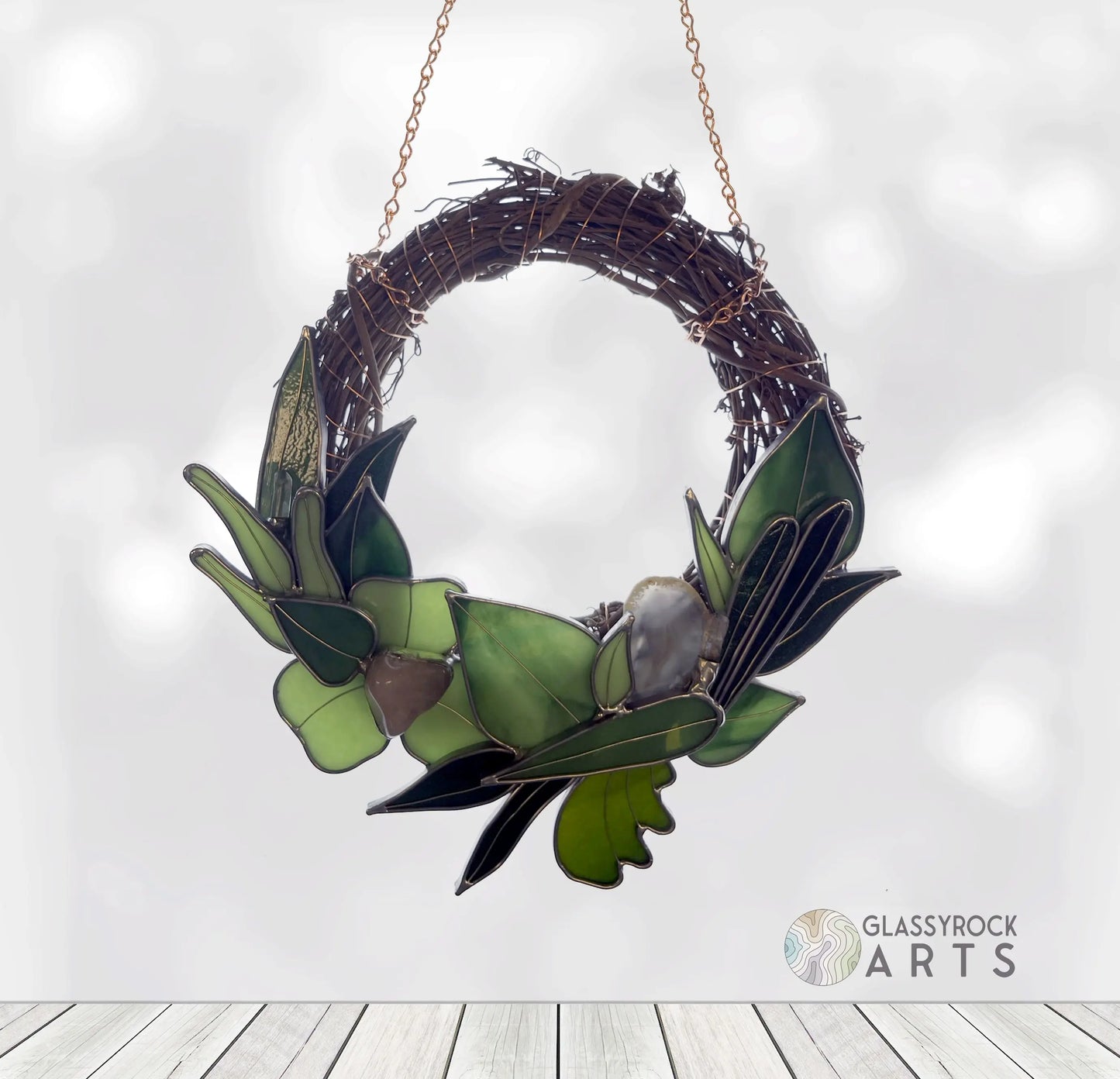 A Handmade Botanical Stained Glass Wreath features shades of green glass leaves and brown vines, hanging by a chain against a softly blurred background. The Glassyrock Arts logo appears at the bottom.