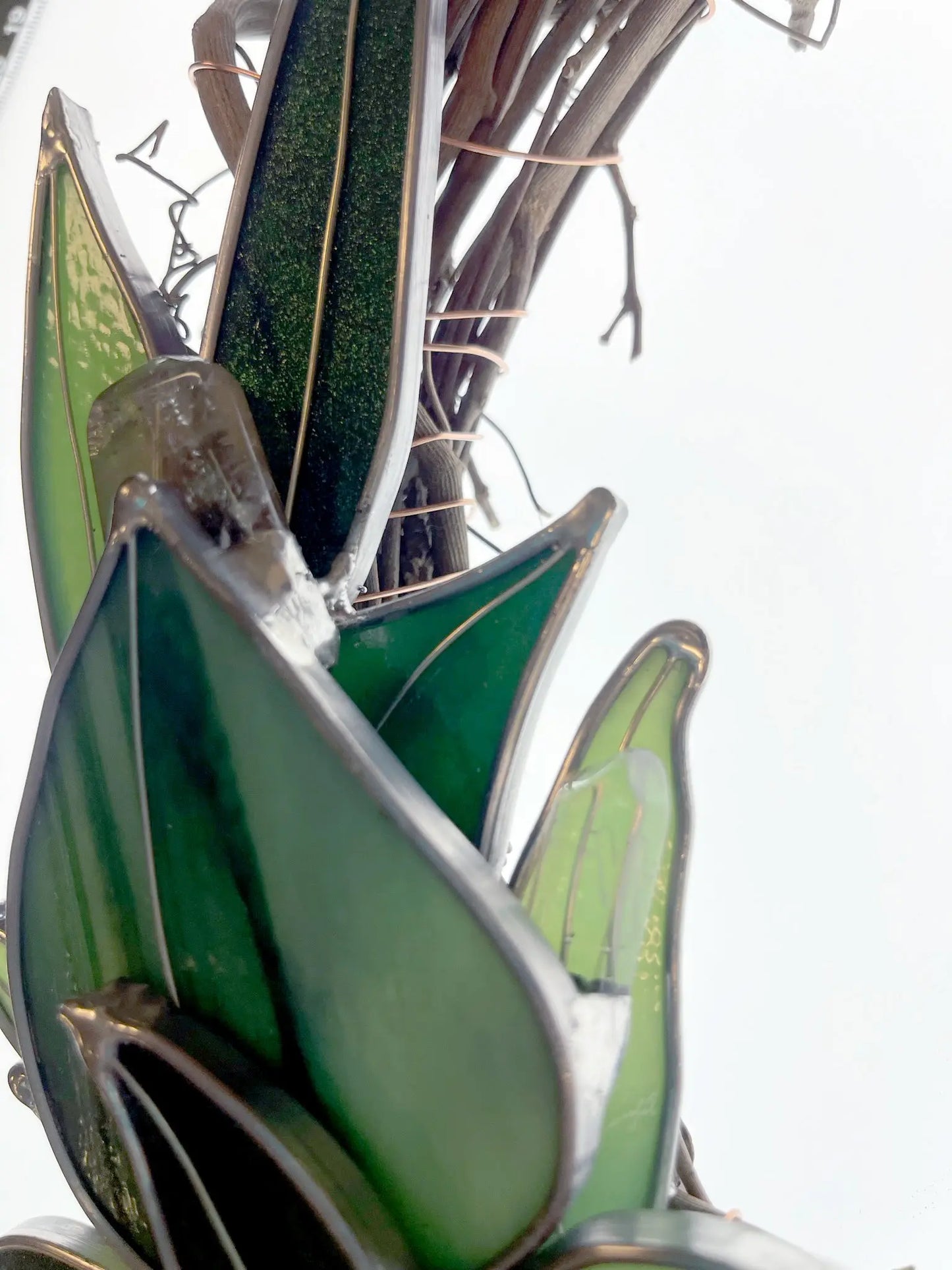 View from below of the Handmade Botanical Stained Glass Wreath: green glass pieces form leaf shapes outlined in metal against a light background, resembling a botanical art installation with twisted branches.