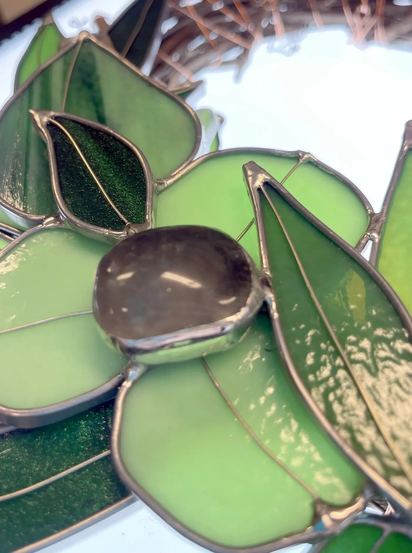A close-up of the Handmade Botanical Stained Glass Wreath reveals green leaves with a glossy black stone center on a light background. Intricate metalwork outlines the leaves, showcasing exquisite craftsmanship and timeless beauty.