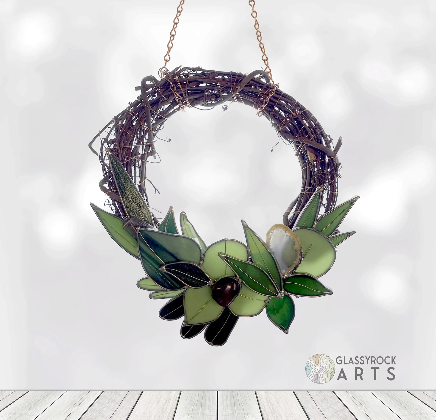 A Handmade Botanical Stained Glass Wreath with glass leaves and olives in various green shades hangs on a chain against a white backdrop, featuring the Glassyrock Arts logo in the corner.