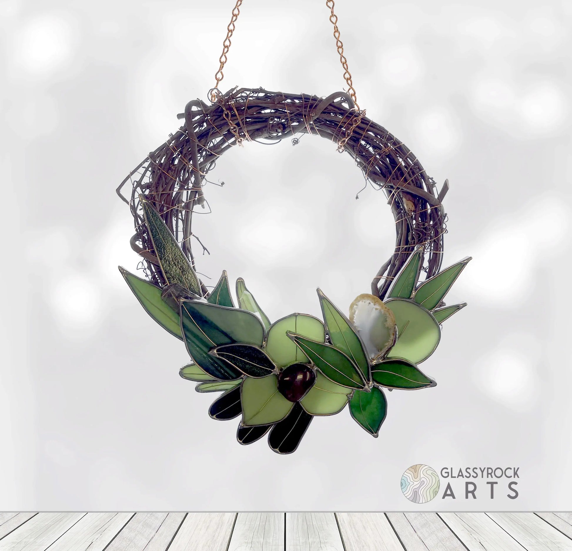 A Handmade Botanical Stained Glass Wreath with glass leaves and olives in various green shades hangs on a chain against a white backdrop, featuring the Glassyrock Arts logo in the corner.