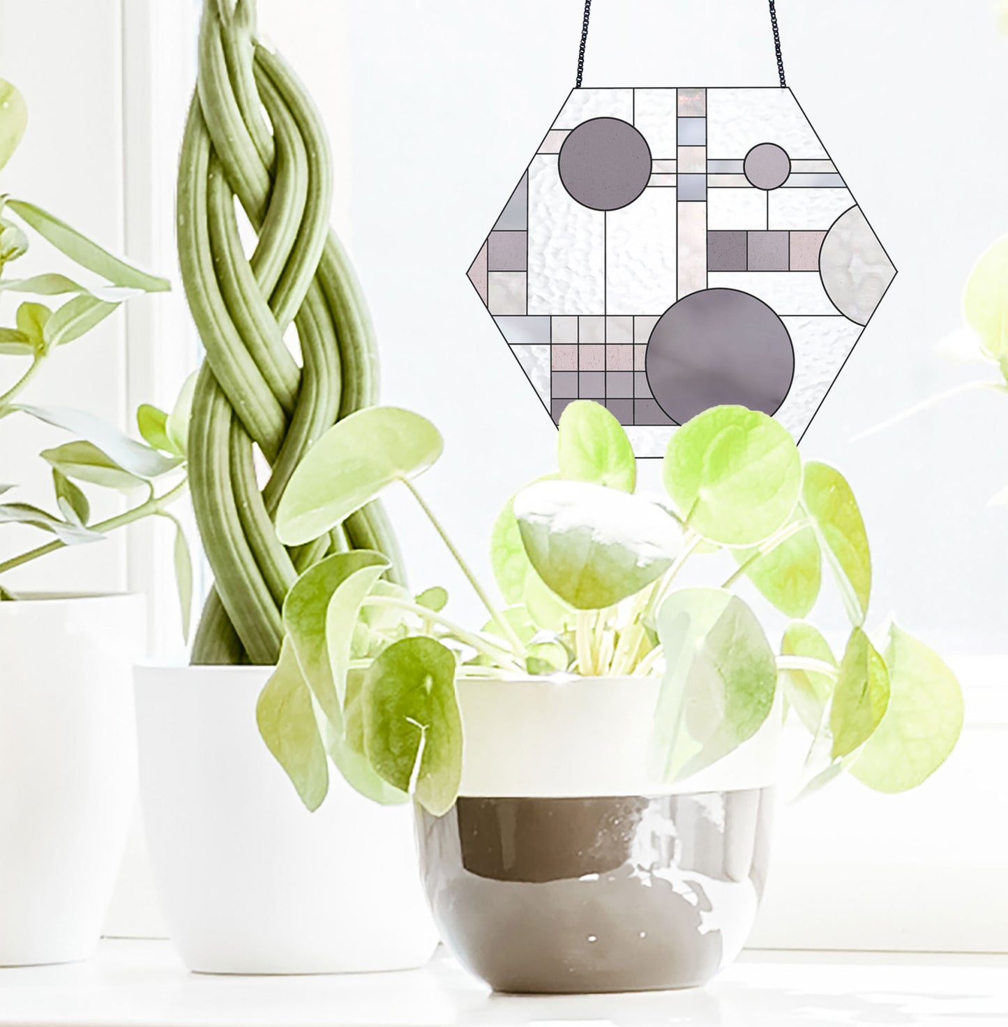 Hexagonal stained glass piece with geometric patterns hangs near a plant in a white pot. The background features a twisted plant in another pot and a bright window, creating a light, airy ambiance.