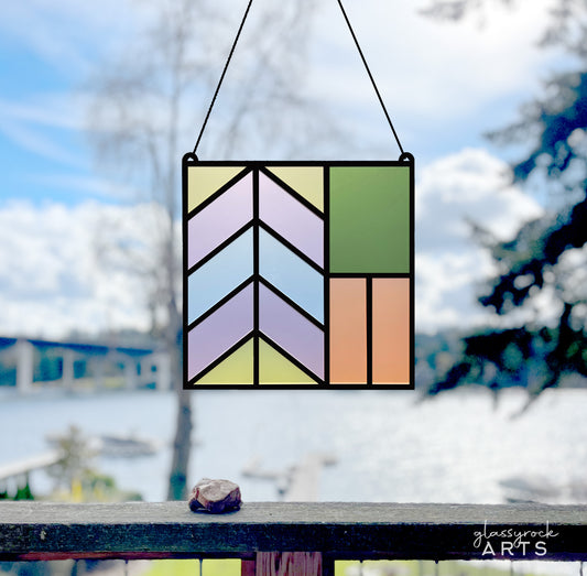 A colorful geometric stained glass panel made from laser-cut acrylic and hung in front of a lake scene with blue sky and large trees.