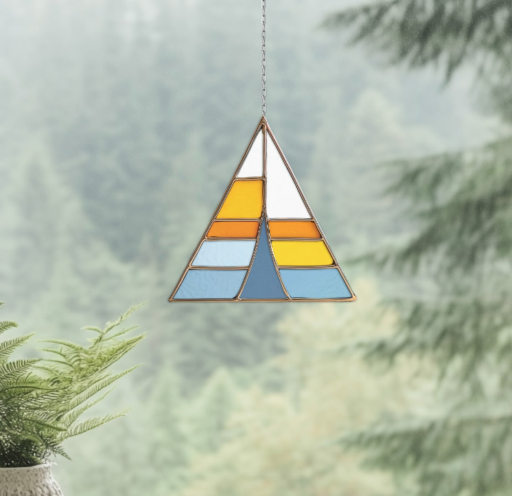 Stained glass panel with geometric design hangs in a sunlit window. A plant with large leaves and a textured pillow are placed on a cozy seating area next to the window. Trees are visible outside.