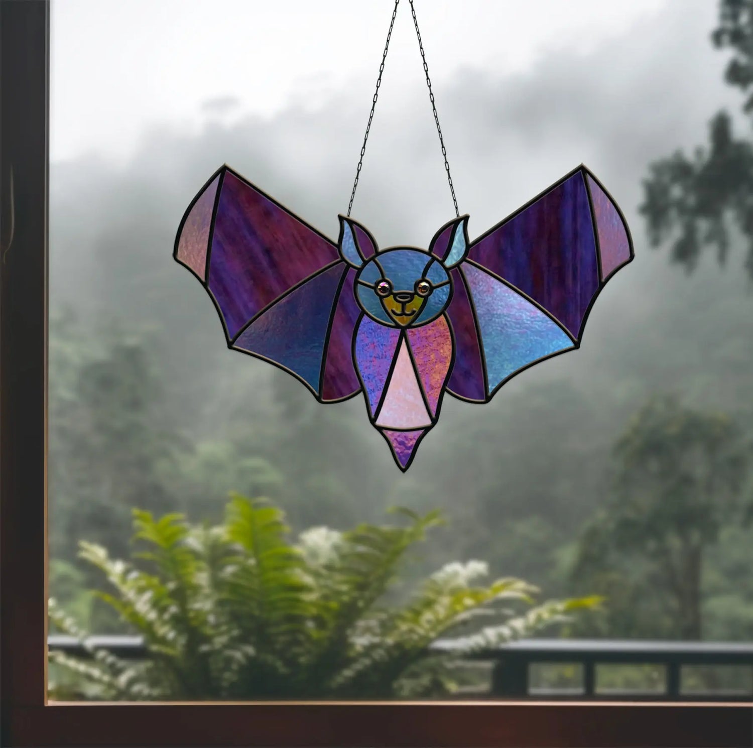 Stained glass bat with purple hues hangs in a window, casting light patterns. Behind it, a view of misty trees and ferns creates a serene atmosphere.