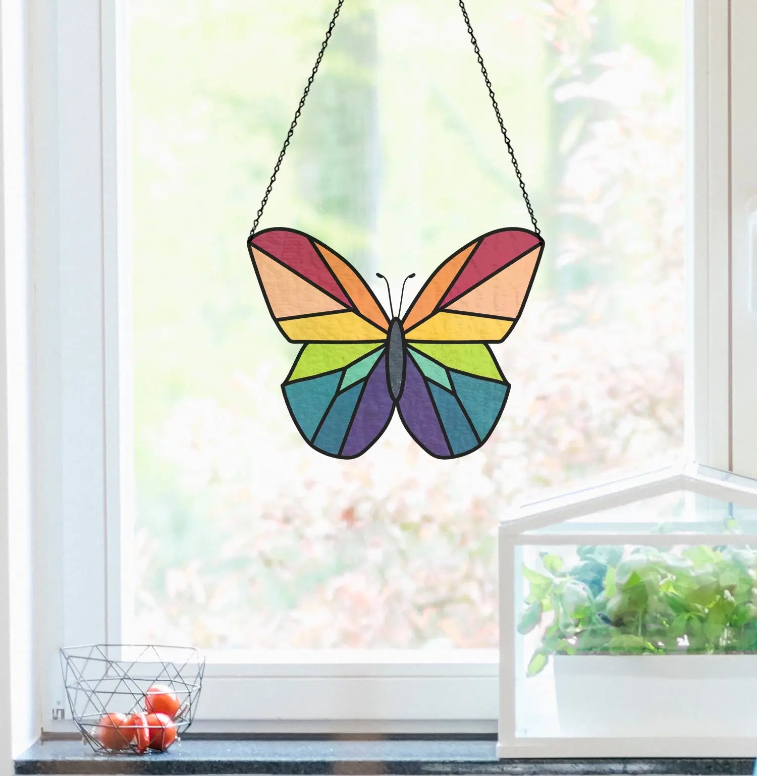 Stained glass butterfly with rainbow colors hanging in a window. A small basket of red tomatoes and a glass plant case with greenery are on the windowsill. Bright natural light filters through.