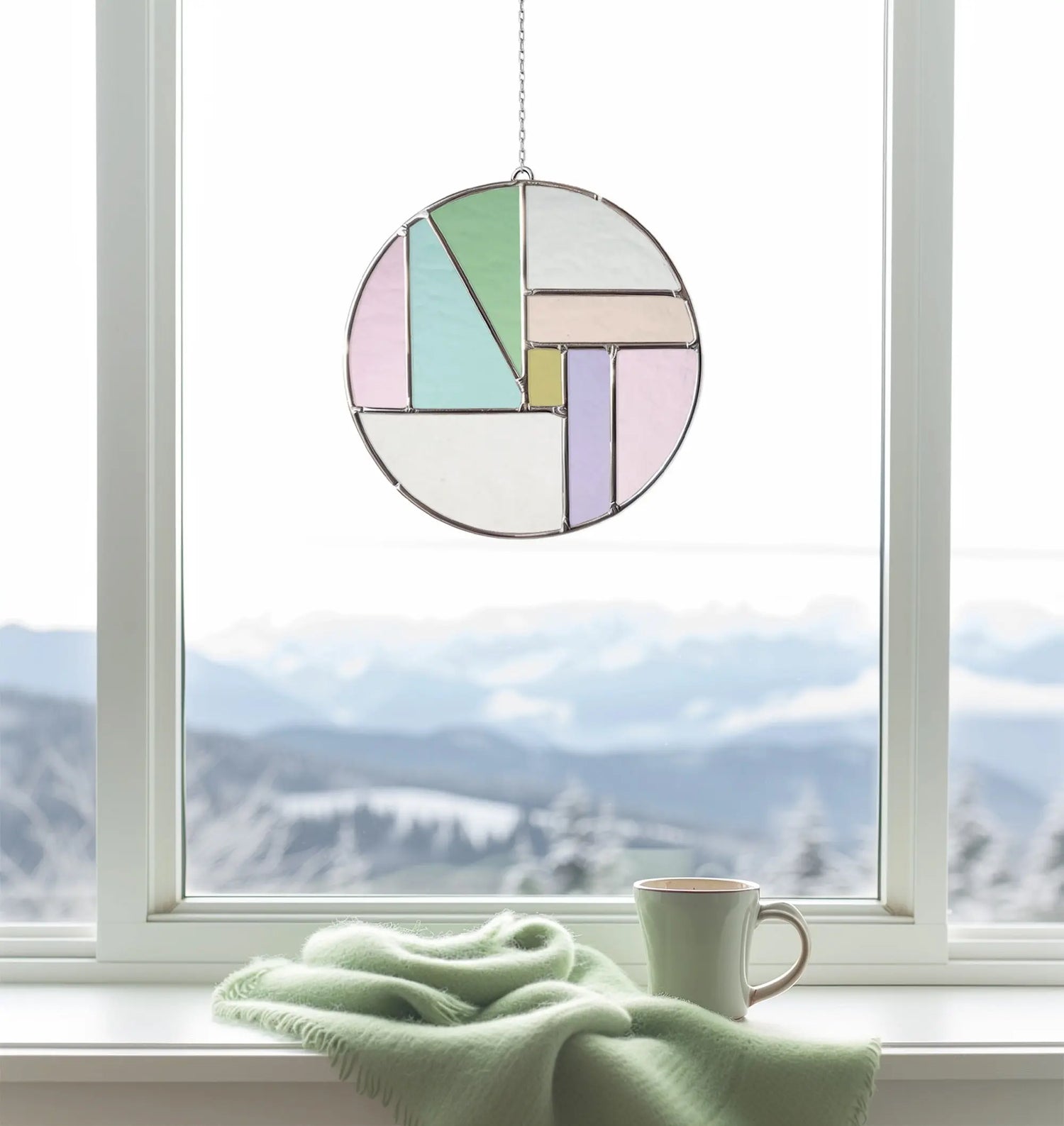 A round stained glass ornament with pastel geometric shapes hangs in front of a window. A green blanket and a mug are on the windowsill. Snowy mountains are visible in the background.