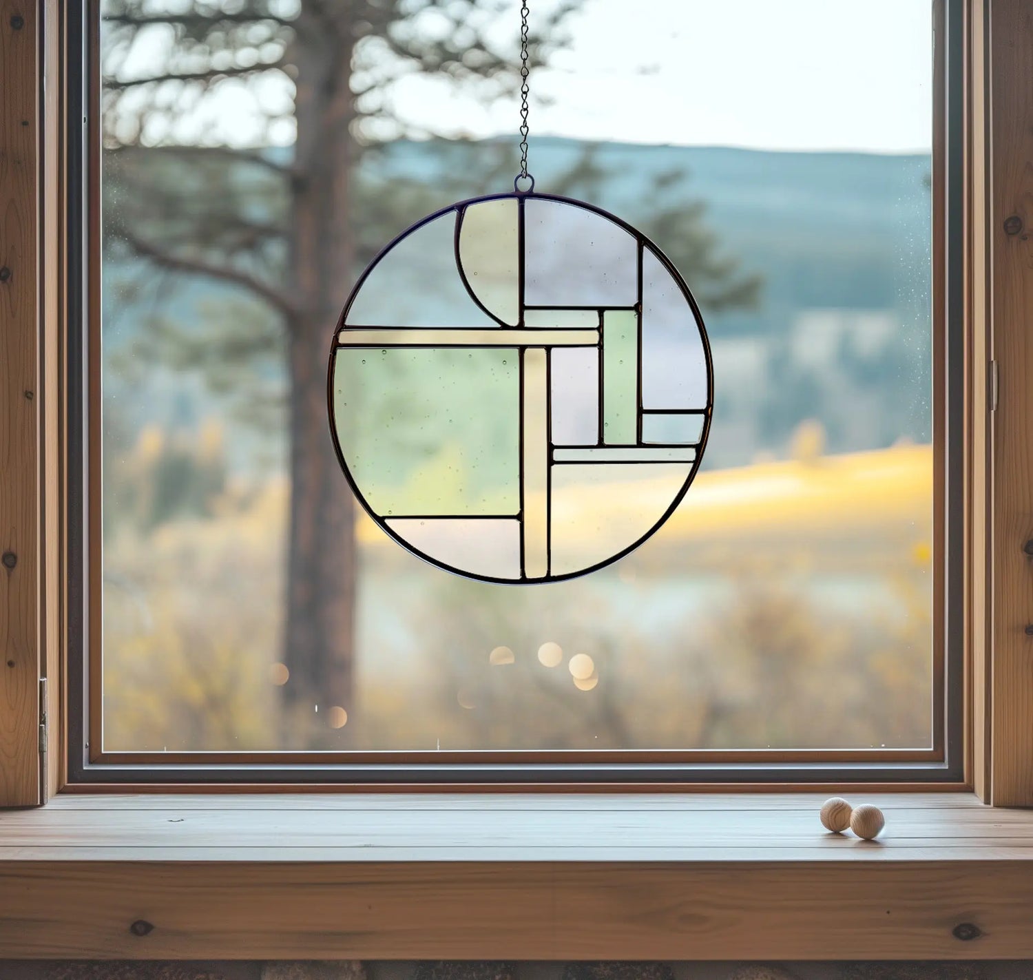 A round, geometric stained glass decoration hangs in a wooden window frame, with a view of a blurred, sunlit landscape featuring trees and hills in the background. A small wooden object sits on the windowsill.