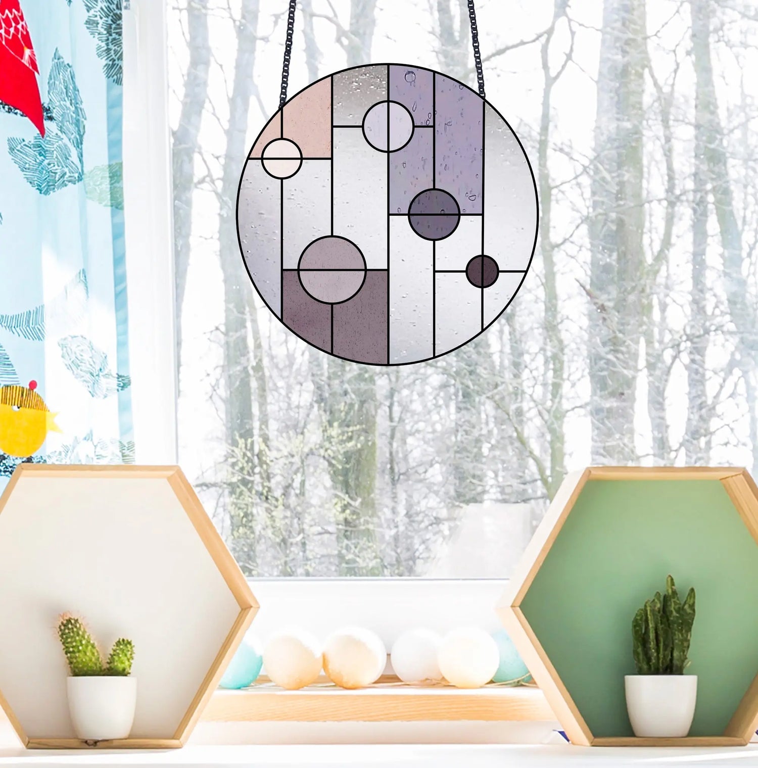 A circular stained glass piece with geometric shapes hangs in front of a snowy window. Below, two hexagonal shelves hold small cacti and decorative objects.