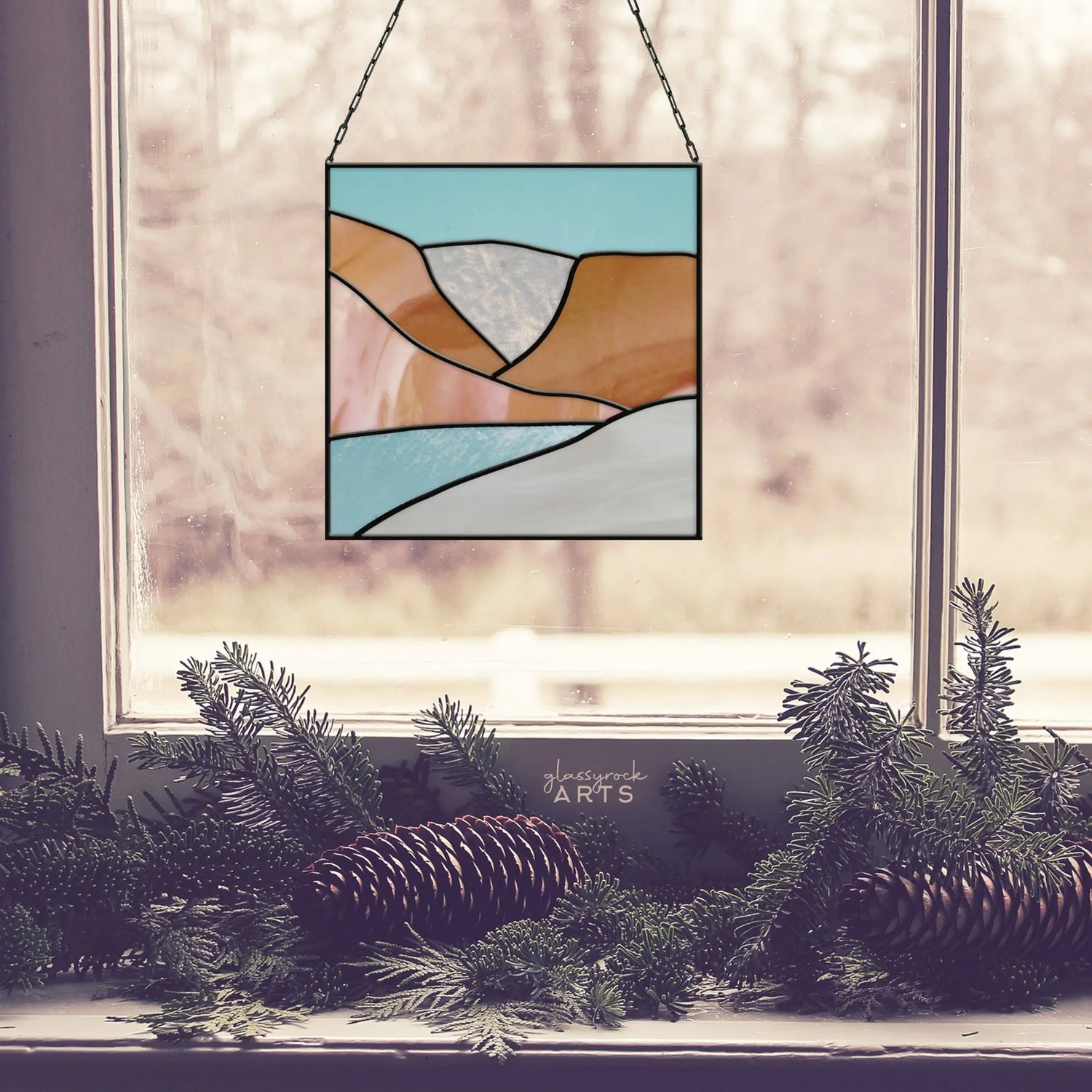 Stained glass artwork featuring a landscape hangs in a window. The design includes blue, orange, and clear sections. Greenery with pinecones is placed on the windowsill, and a blurry background is visible outside.