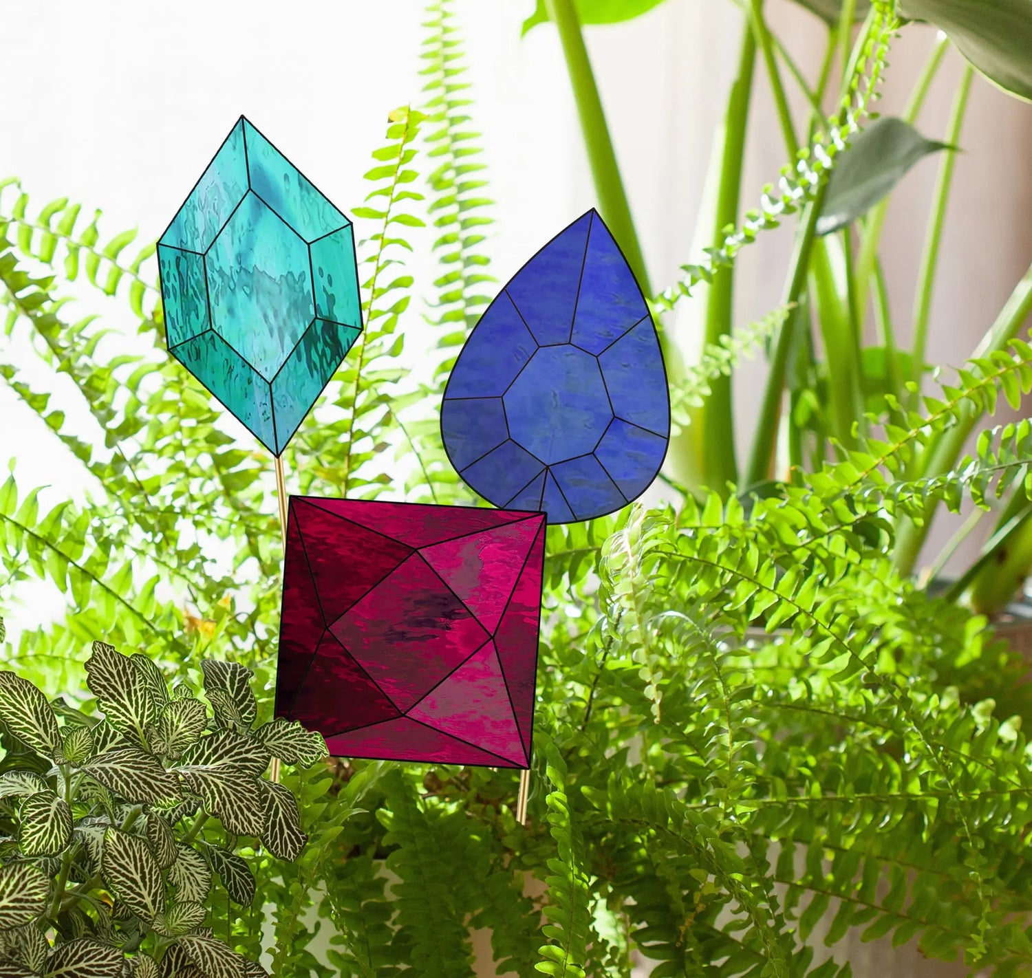 Three stained glass garden stakes in blue, purple, and red gem shapes are placed among lush green plants and ferns indoors, creating a vibrant and artistic display. Sunlight filters through, highlighting the colors.