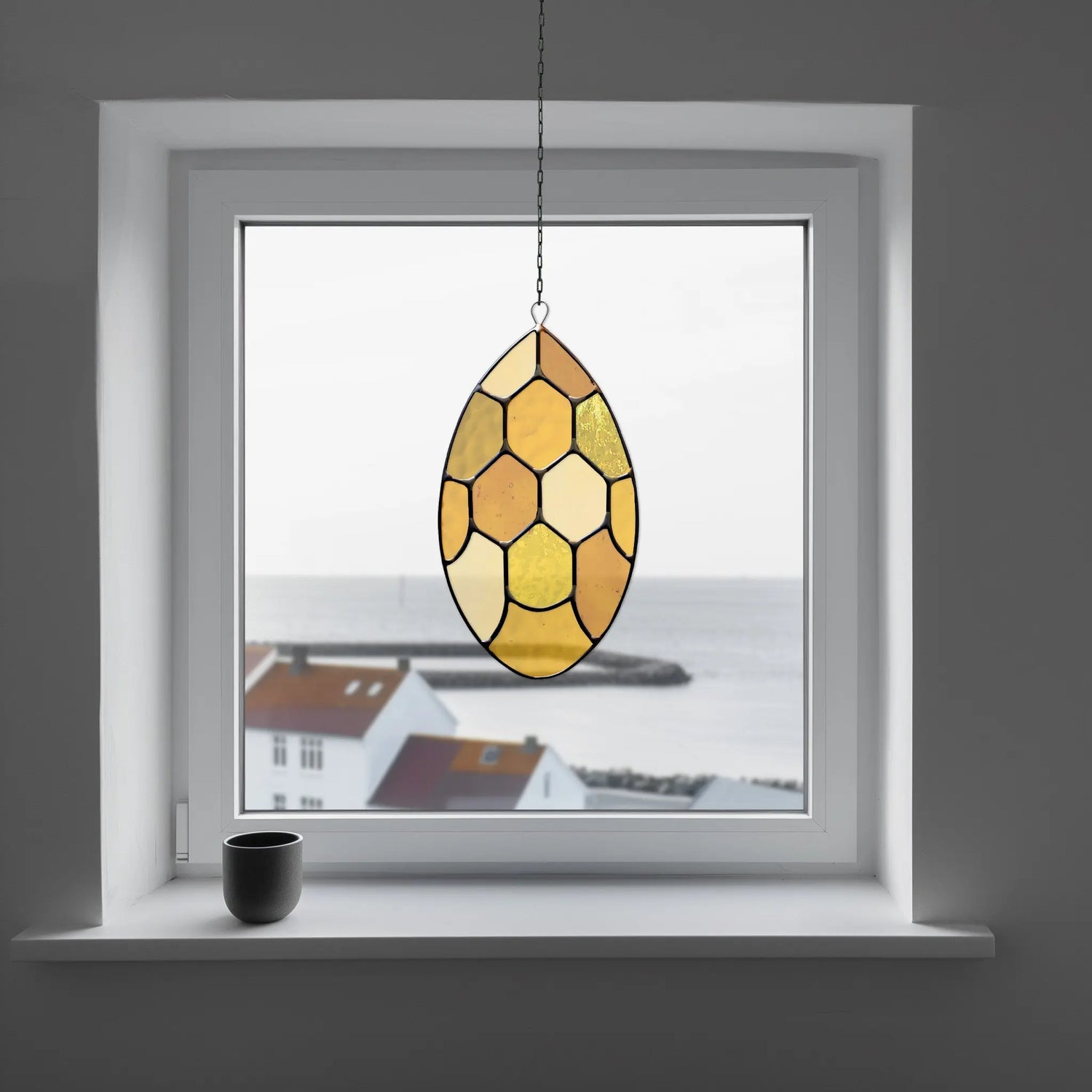 Sunlit, hexagonal-patterned stained glass hangs in a window. Beyond, a coastal view with white houses and a calm sea. A black mug sits on the windowsill, adding a minimalistic touch.