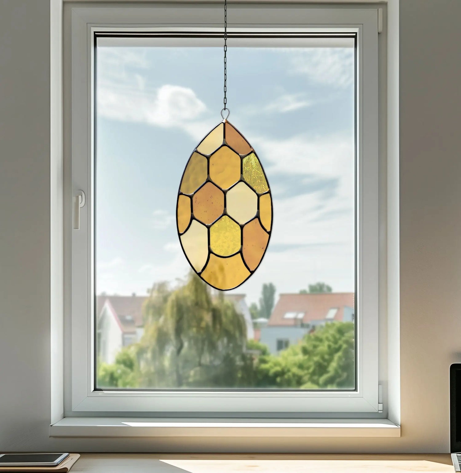 A stained glass oval sun catcher with shades of orange and yellow hangs by a chain in a window. Outside, a clear blue sky and houses with red roofs are visible.