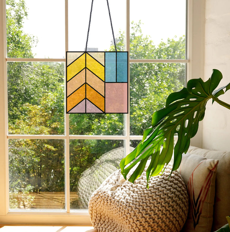 Stained glass panel with geometric design hangs in a sunlit window. A plant with large leaves and a textured pillow are placed on a cozy seating area next to the window. Trees are visible outside.