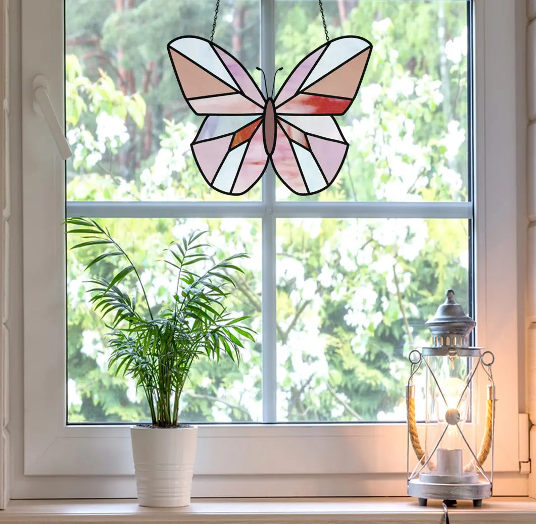 Stained glass butterfly hanging in a window with a view of lush green trees and white flowers outside. A potted plant and lantern sit on the windowsill.