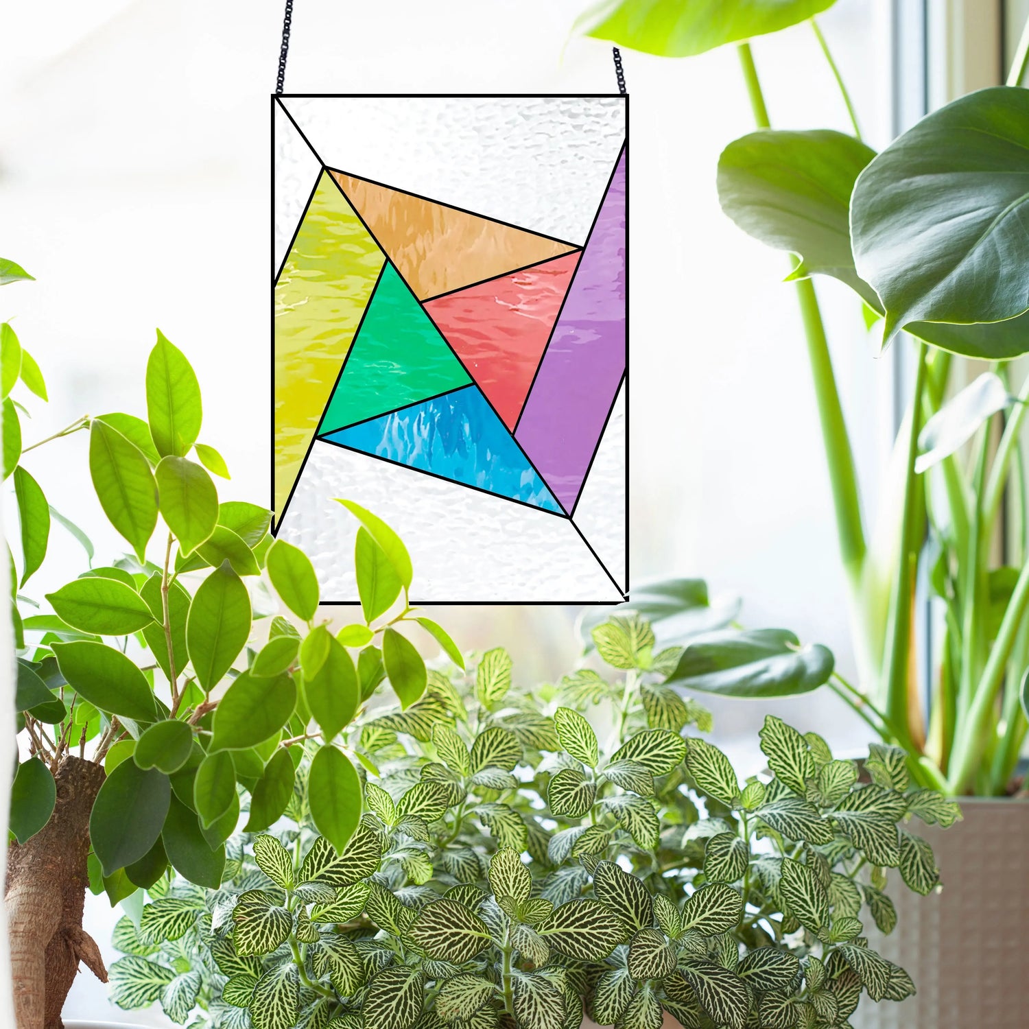 A colorful geometric stained glass panel hangs amidst lush green houseplants by a sunlit window, casting vibrant reflections.