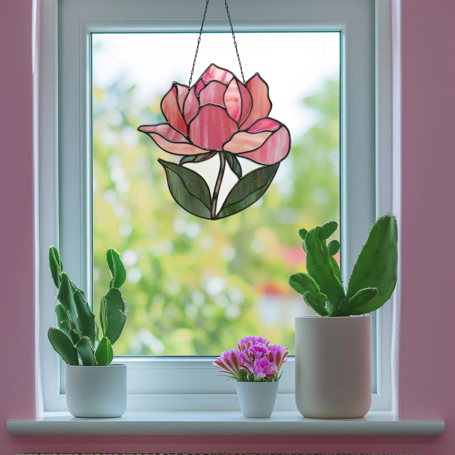 Two cacti and a small flowering plant sit on a windowsill. A stained glass ornament depicting a pink flower hangs in the window, with a blurred green and pink background visible outside.