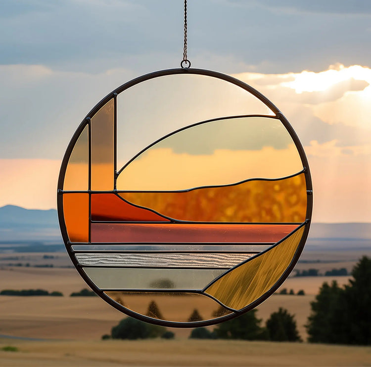 A circular stained glass panel hangs against a landscape backdrop. It features geometric patterns in warm tones, reflecting a serene sunset over fields with distant mountains and trees beneath a cloudy sky.