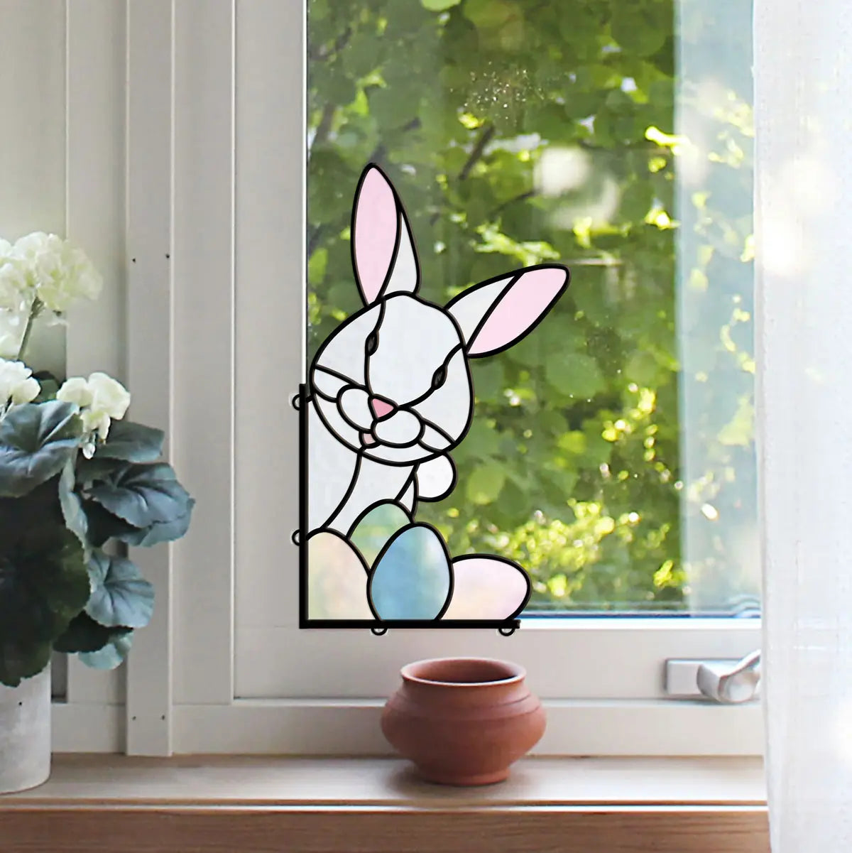 Stained glass window decoration featuring a bunny with pink ears and pastel-colored eggs, placed by a window with green foliage outside. A terracotta pot and white flowers adorn the windowsill.