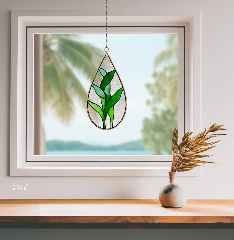 A teardrop-shaped stained glass with green leaf design hangs in front of a window overlooking a blurry tropical scene. A small vase with dried plants sits on the windowsill.